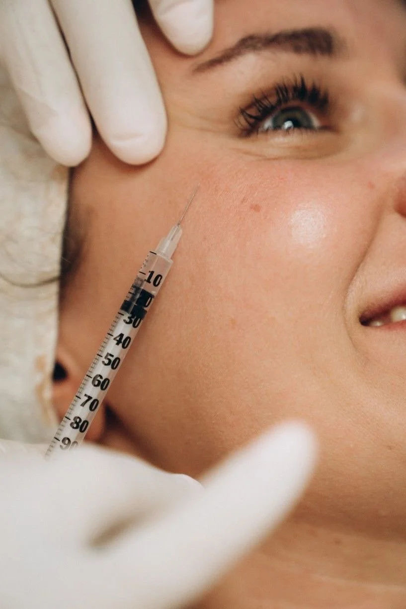 A person receiving a cosmetic injection in their cheek, with a healthcare professional wearing gloves preparing the syringe.