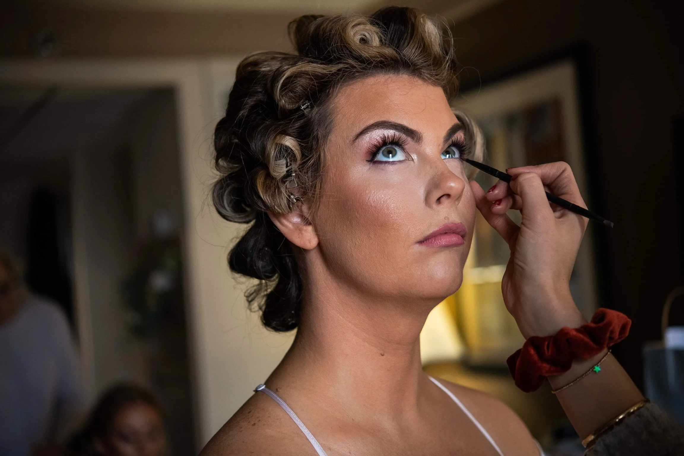 Woman with styled hair getting her makeup done, looking upward as makeup artist applies eyeshadow with a brush.