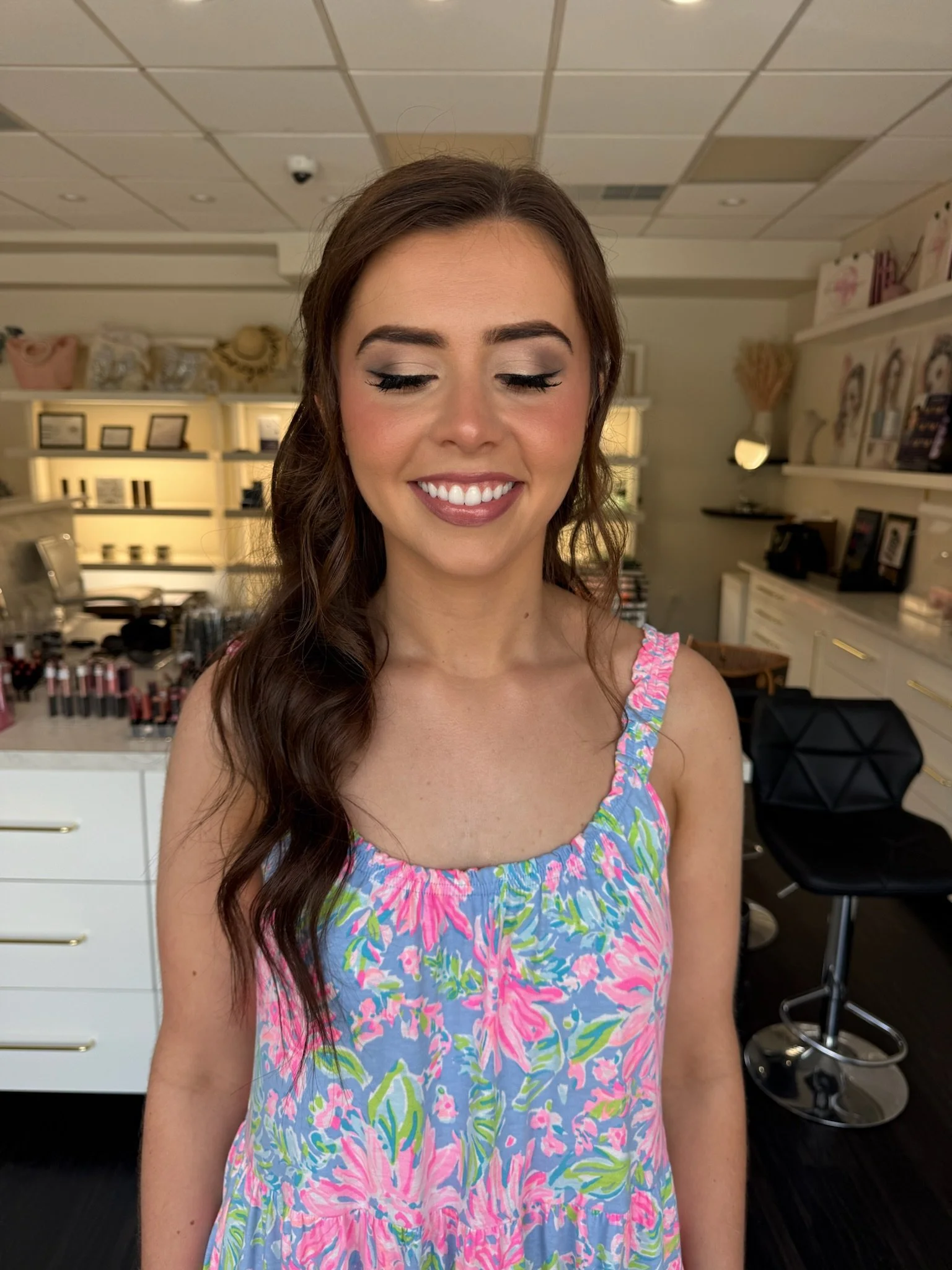 A young woman with long brown wavy hair, closed eyes, and a bright smile, wearing a colorful sleeveless floral dress, standing inside a makeup store with shelves of makeup products and framed photos in the background.