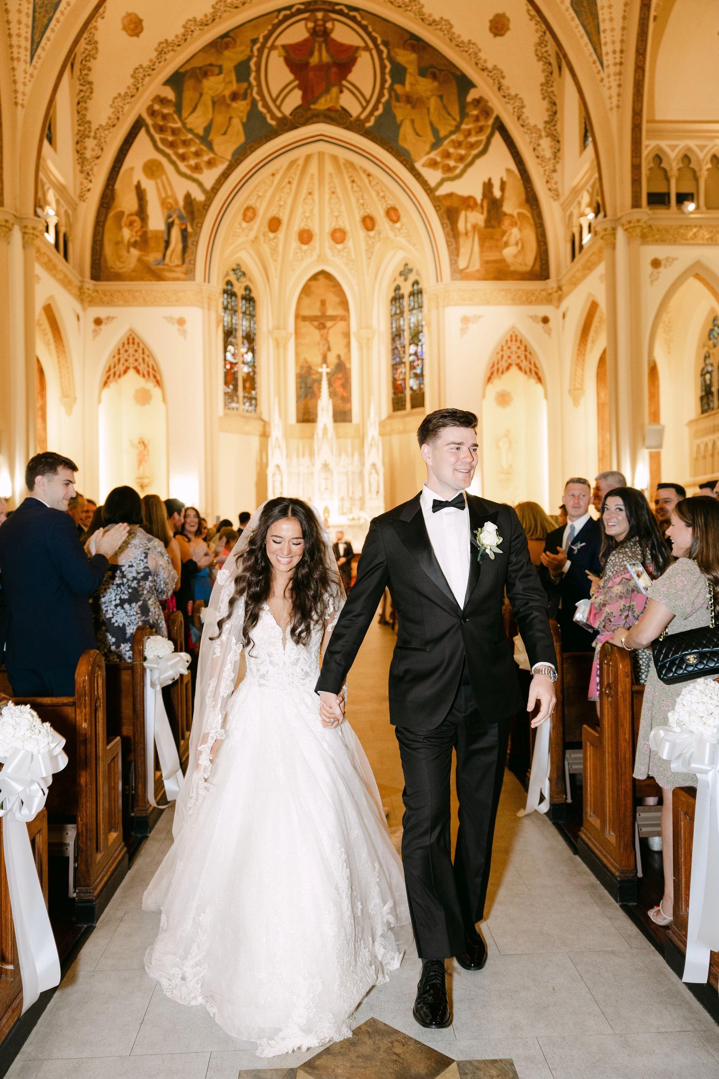 A newlywed couple walks down the aisle of a church, holding hands and smiling, surrounded by wedding guests in a decorated cathedral with religious artwork.