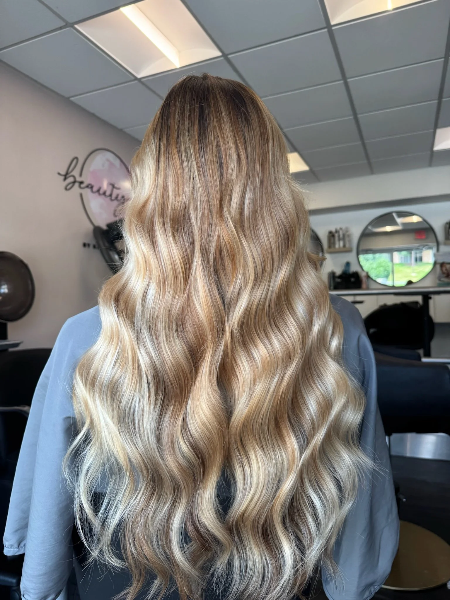 Back view of a woman with long, wavy blonde hair in a salon.