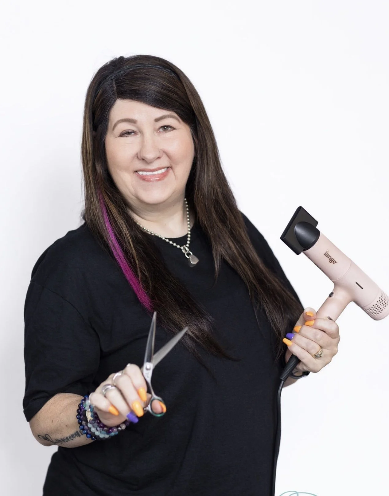 A woman with long brown hair with pink highlights, wearing a black shirt, a pearl necklace, and a black headband, smiling at the camera against a white background.