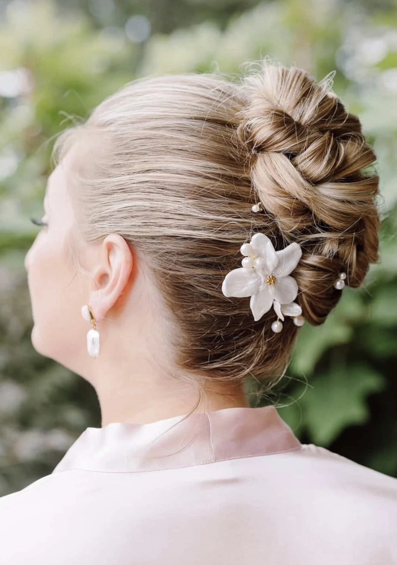 Back view of a woman with a braided updo hairstyle decorated with white flowers and pearls, wearing pearl earrings and a light-colored satin top. Green foliage in the background.