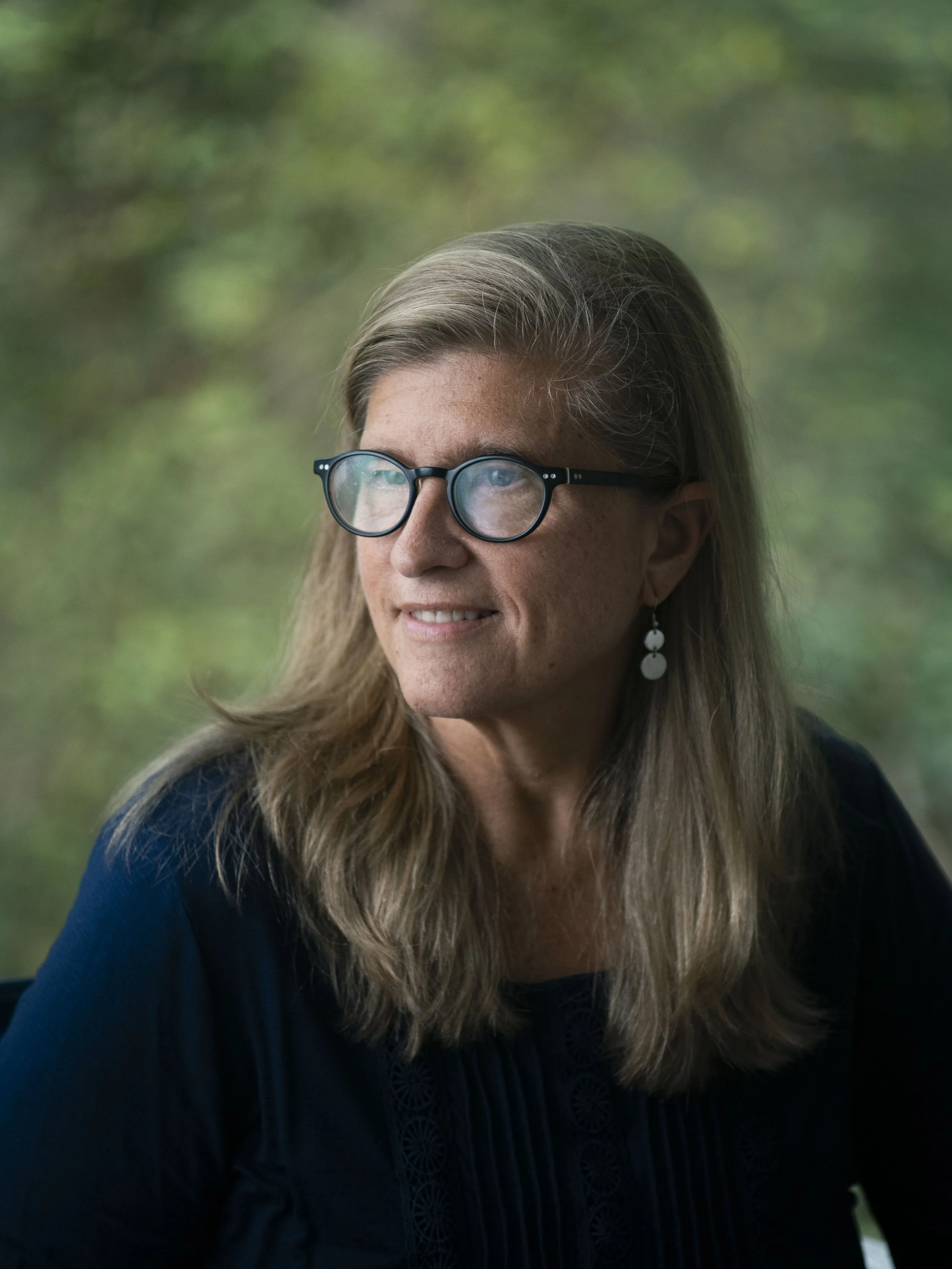 A woman with long, wavy gray hair wearing glasses and earrings, looking to the side against a blurred green outdoor background.