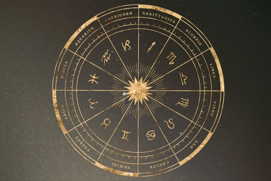 A gold-colored zodiac wheel with the 12 astrological signs, set against a black background. The signs are arranged in a circle with degree markings, and the names of the signs are written in English around the outer edge.