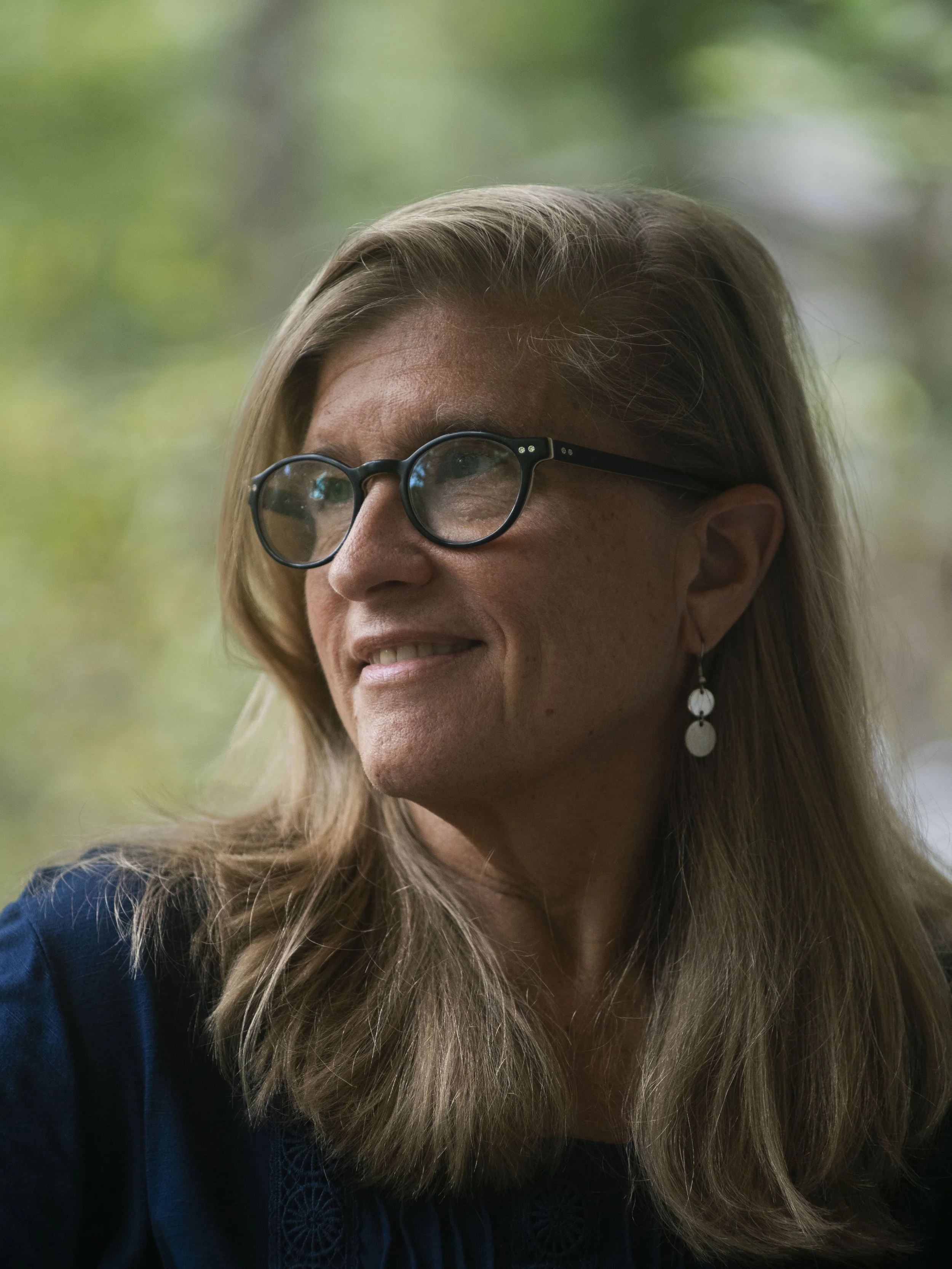 A woman with long hair, glasses, and earrings, smiling and looking to the side with a blurred green background.