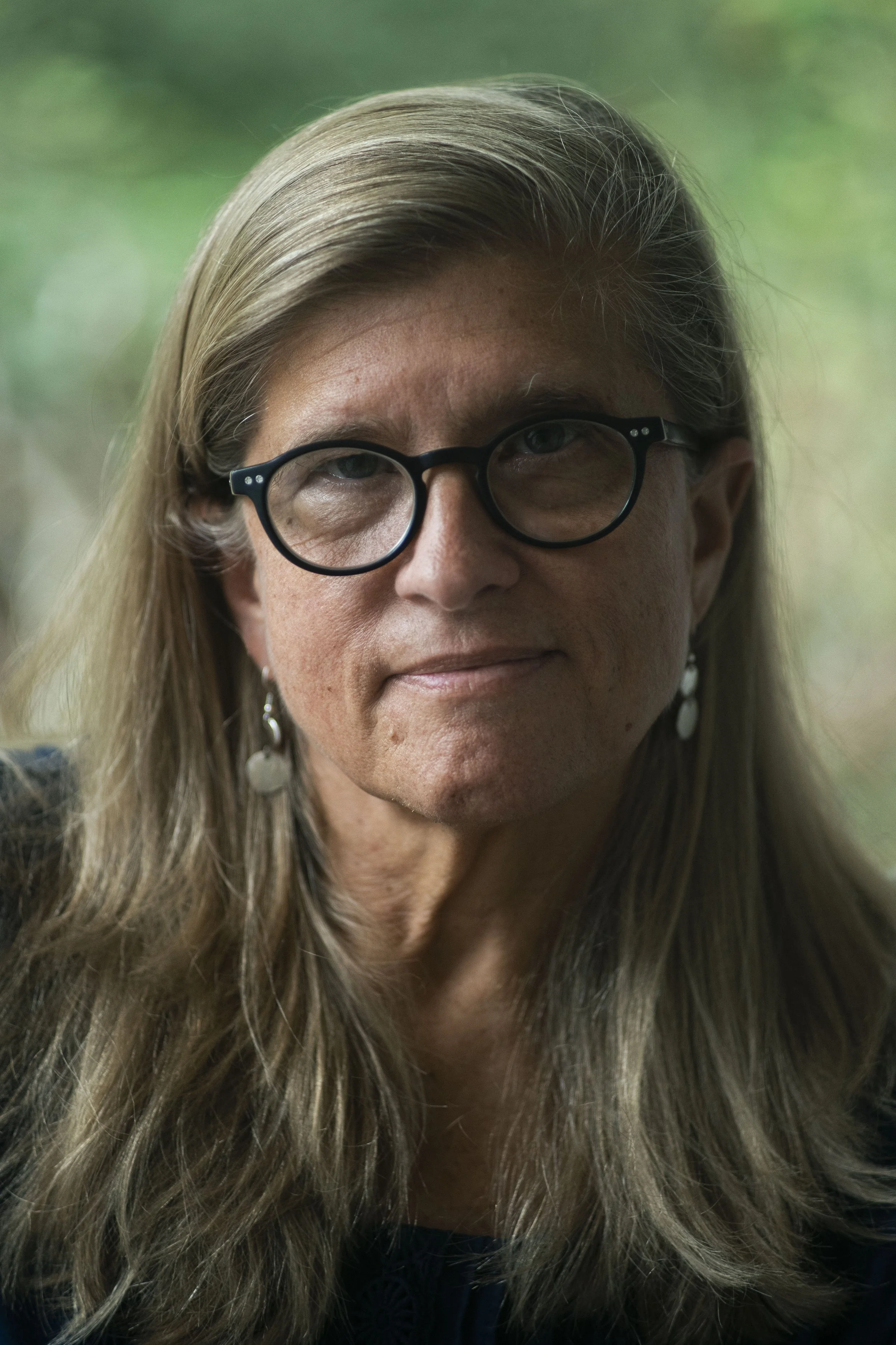 Close-up of a middle-aged woman with long, straight blonde hair, wearing black-rimmed glasses, earrings, and a dark top, against a blurred green background.