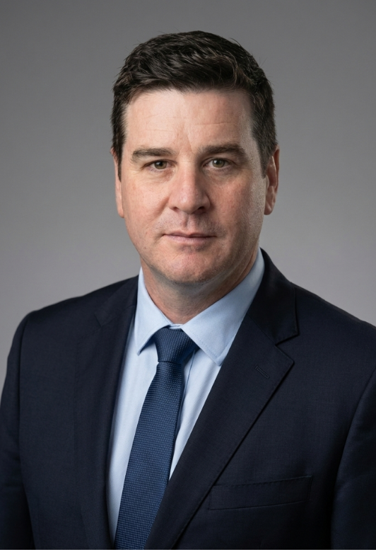 A professional headshot of a Chad Penny, founder of Maqro Strategies in a dark blue suit, light blue dress shirt, and matching blue tie against a neutral gray background.