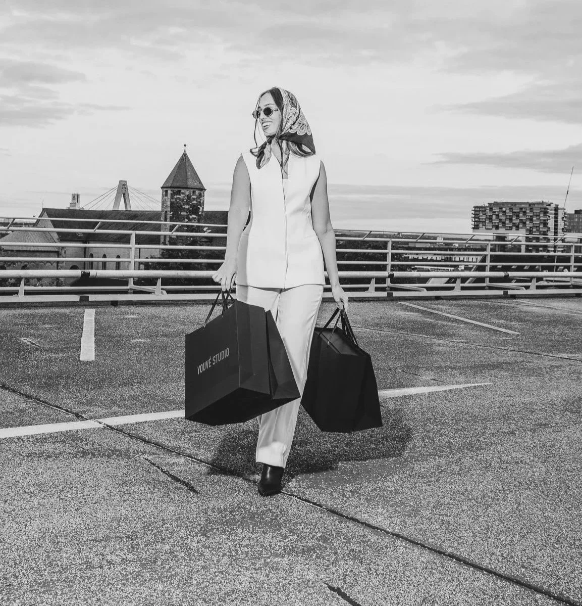 A woman in a stylish outfit holding shopping bags standing on a rooftop parking lot with city buildings and a bridge in the background.