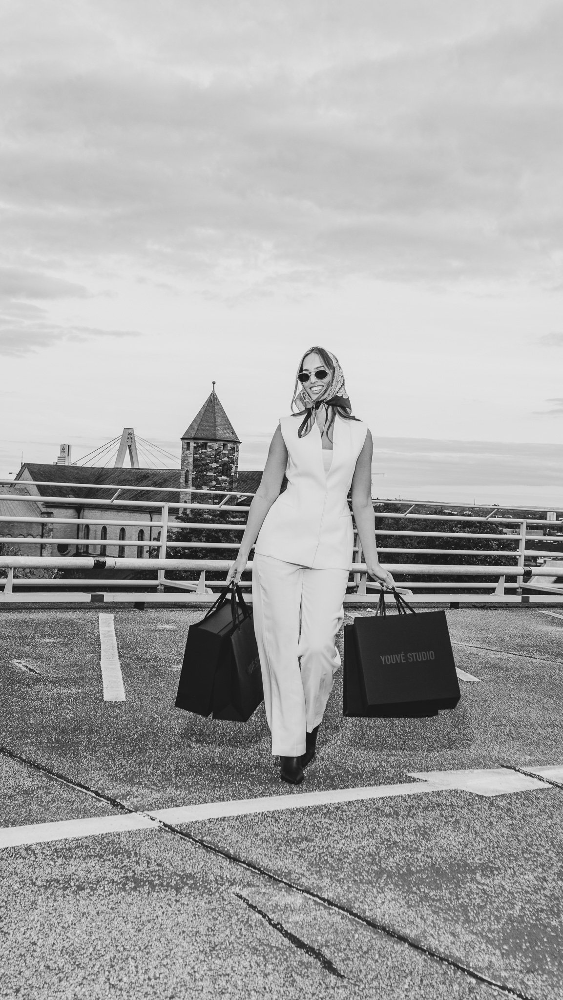 A woman wearing sunglasses and a headscarf, dressed in a white sleeveless suit, walking in a parking lot carrying shopping bags.
