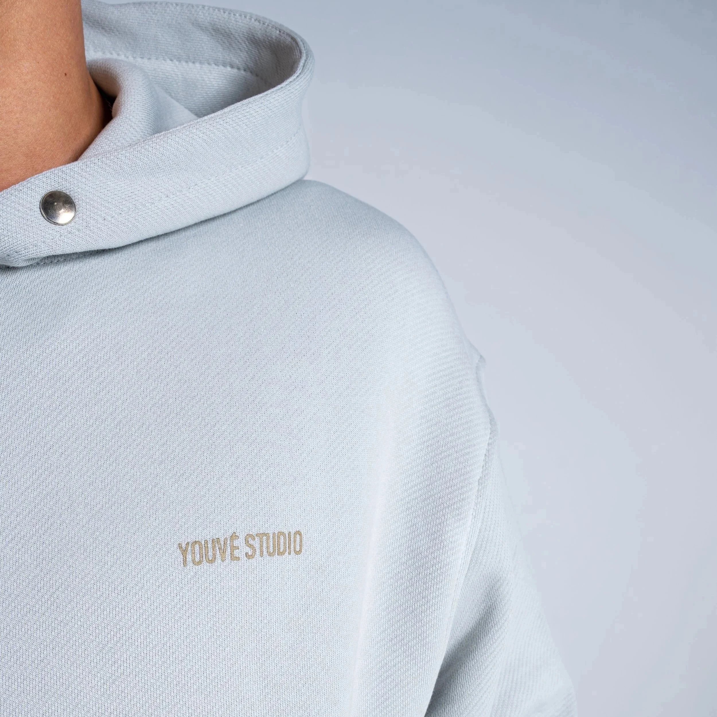 Close-up of a white hoodie with a silver snap button on the collar and faint beige text that says "YOU'VE STUDIO" on the chest.