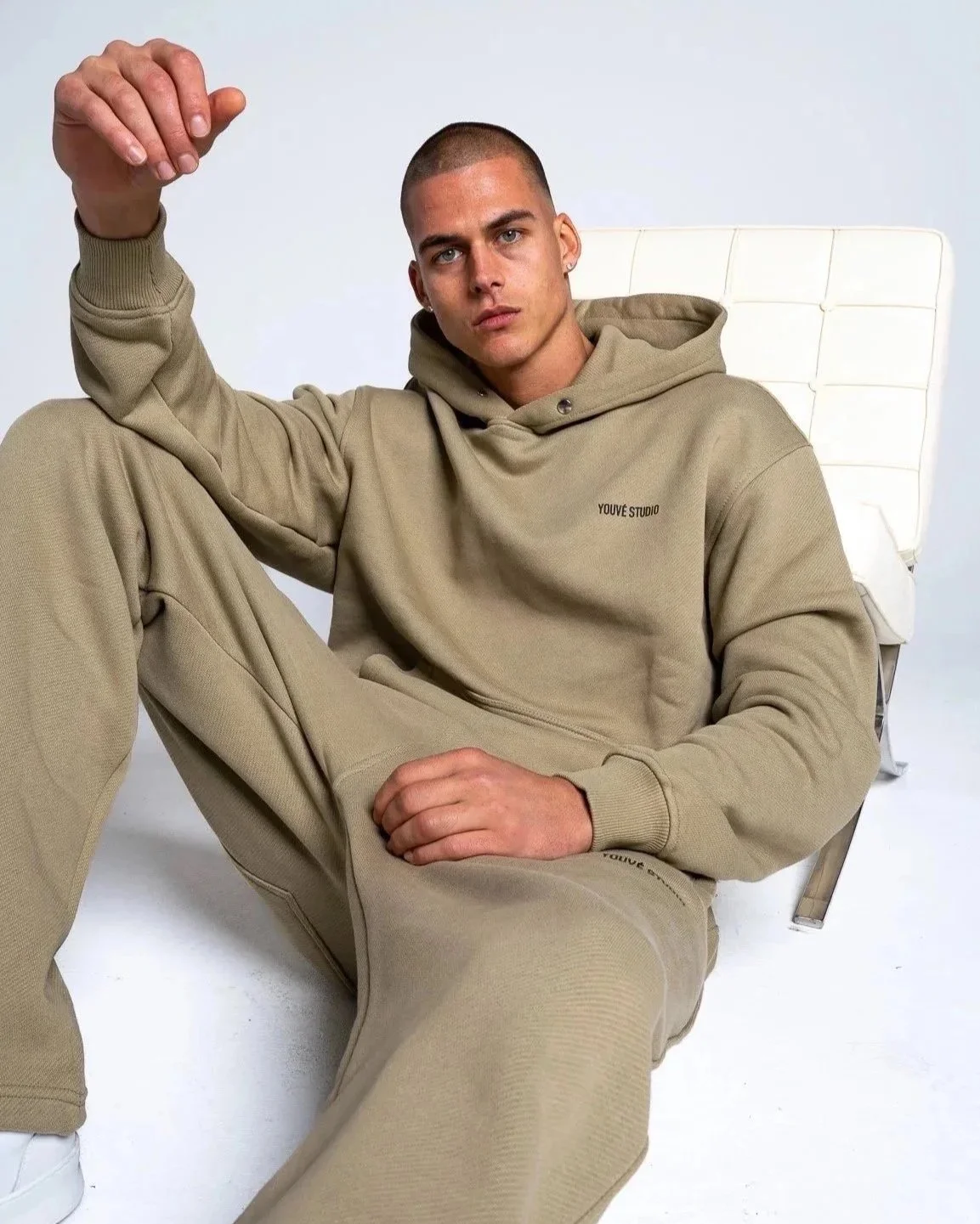 A young man sitting on the floor in a beige hoodie and sweatpants, leaning against a white chair, looking at the camera with a serious expression.
