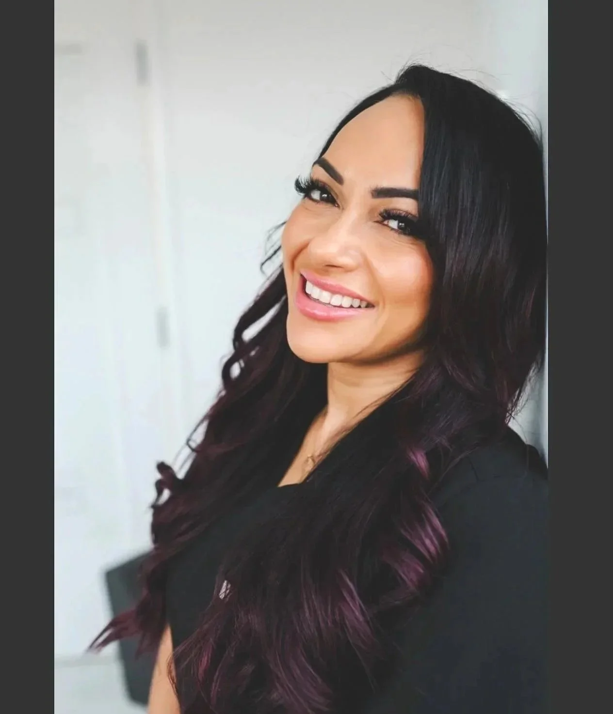 A woman with long dark hair with purple highlights, smiling with white teeth, in a black top, against a plain white background.