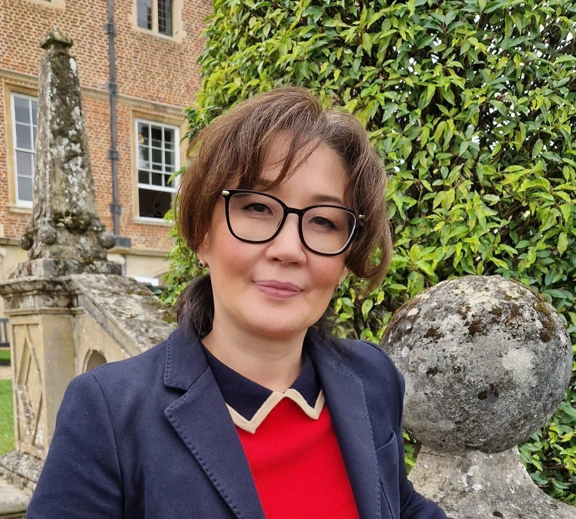 A woman with short brown hair and glasses, wearing a navy blazer, standing outdoors next to Cambridge University College and green hedge in front of a brick building.
