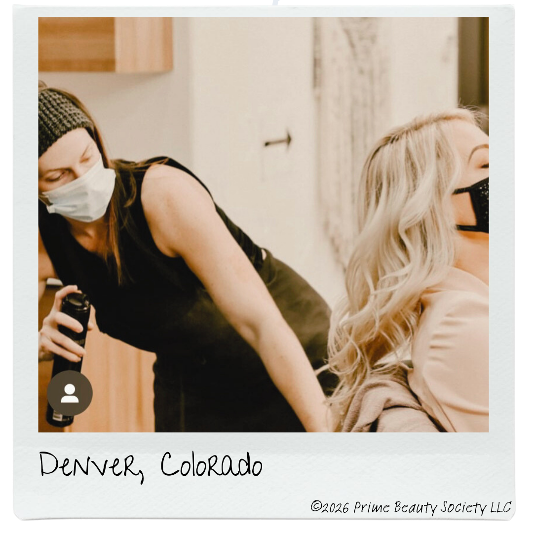 A woman with blonde hair and a face mask is seated while another woman with a face mask and a headband is standing behind her, holding a spray bottle, in a room with a wooden cabinet and light-colored walls, in Denver, Colorado.