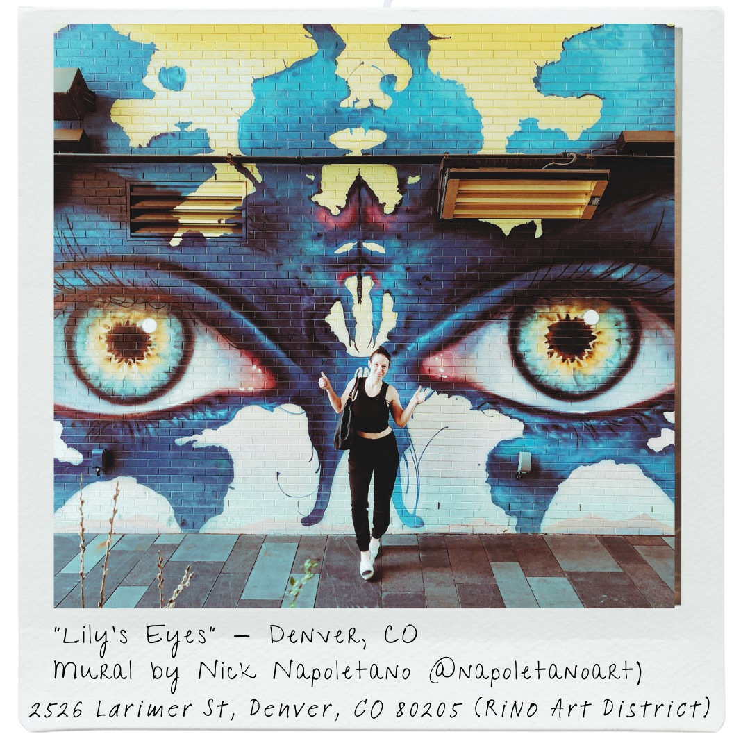 A mural titled 'Lily's Eyes' by Nick Napoletano on a brick wall in Denver, Colorado. The mural features a large pair of realistic, detailed eyes with yellow irises and black pupils, set against abstract blue, purple, and yellow background colors. A woman is standing in front of the mural, gesturing with her hands.