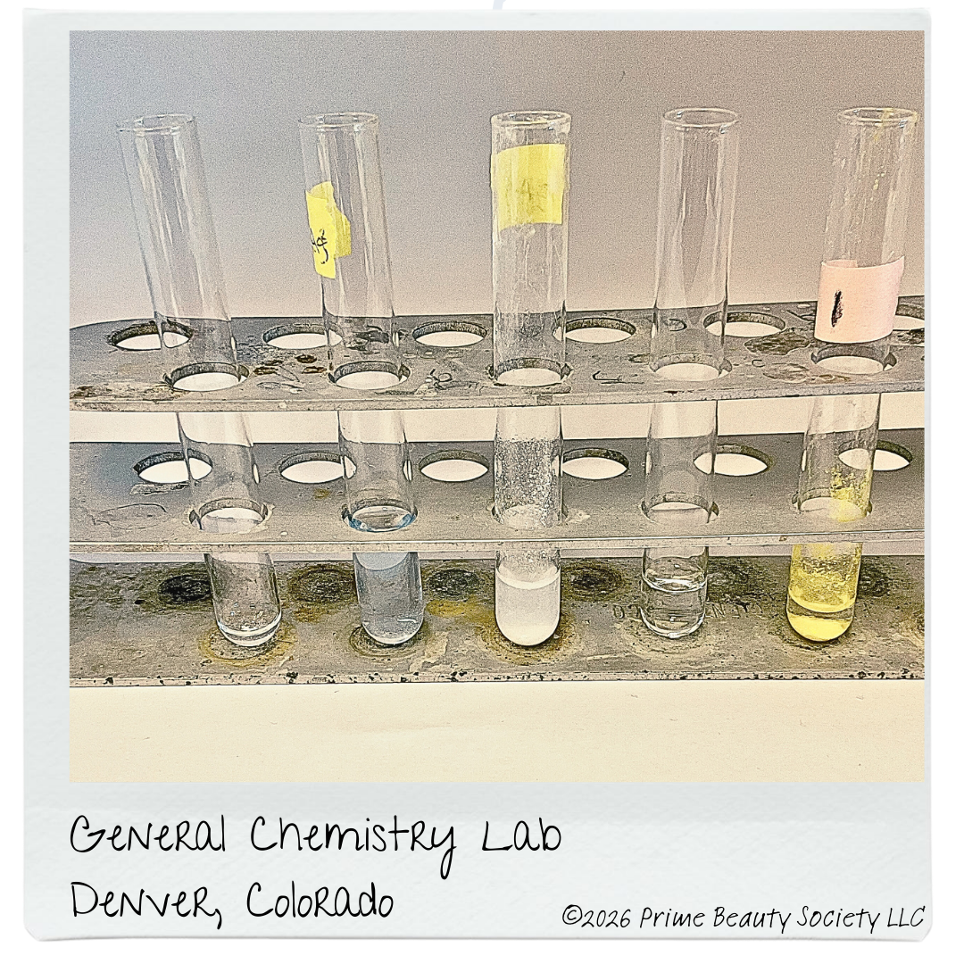 Six test tubes in a metal rack, some with liquids, labeled with different sticky notes, in a general chemistry lab setting. Handwritten note indicates location as Denver, Colorado.