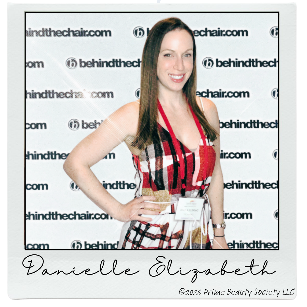 A woman with long brown hair, smiling, wearing a patterned sleeveless dress with red, white, black, and mustard colors, standing in front of a backdrop with repeated 'behindthechair.com' logos. She has a name tag hanging around her neck and is posing with her right hand on her hip.