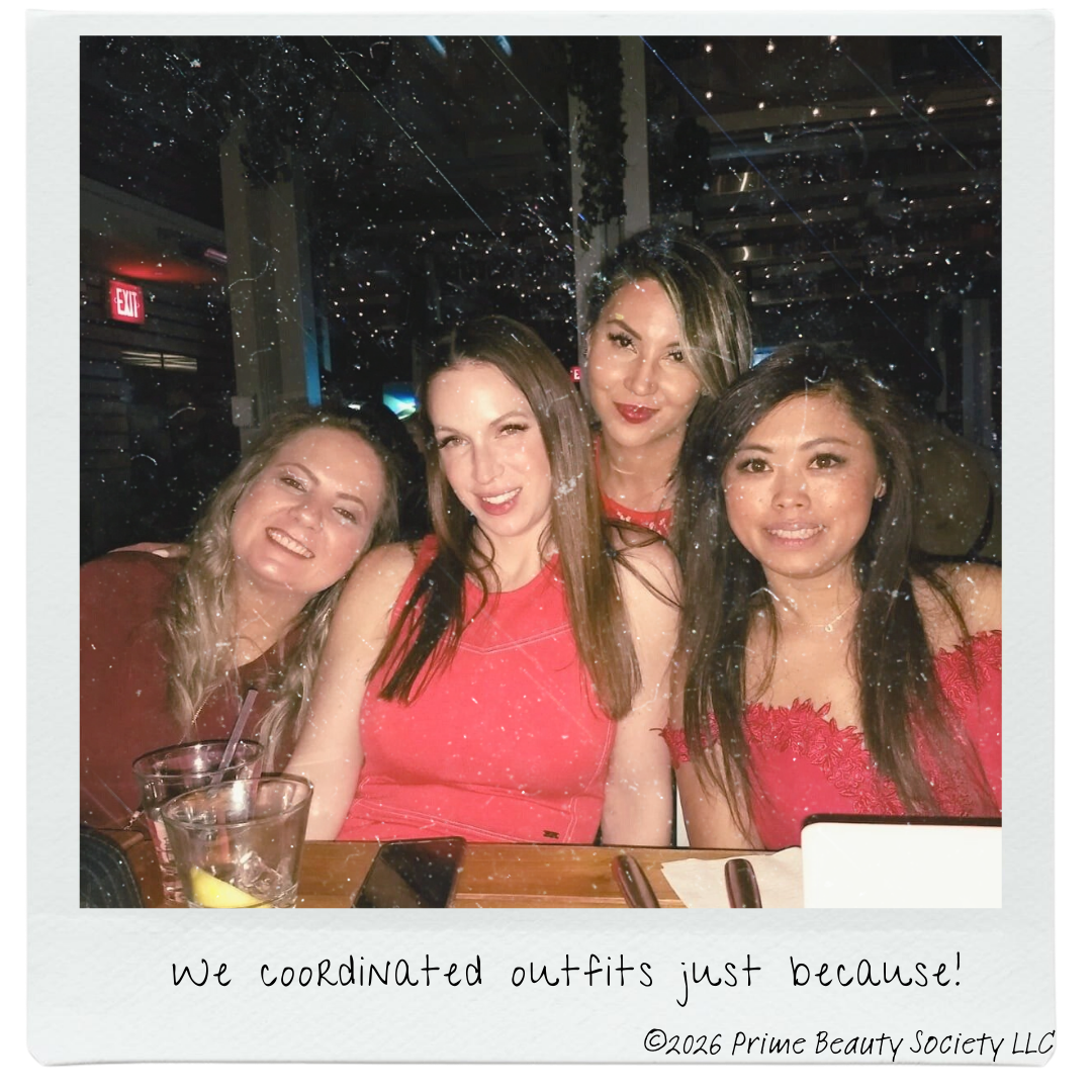 Four women sitting together at a table, dressed in red, smiling at the camera, with a festive atmosphere and sparkly decorations in the background.