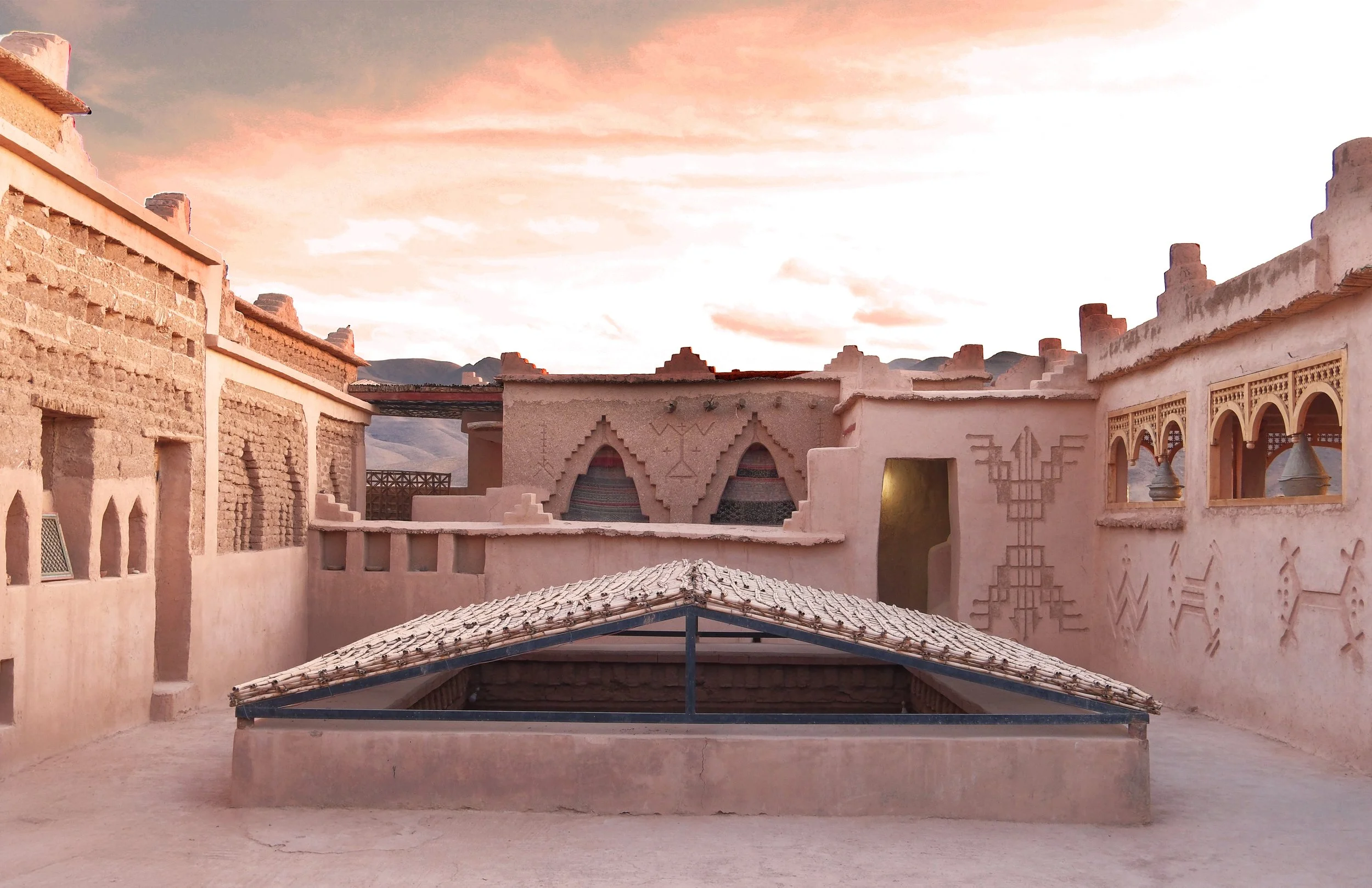 Traditional desert adobe buildings with decorative patterns and small windows, under a pink and orange sunset sky, in a mountainous landscape.