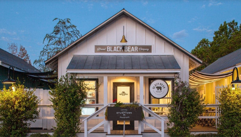 A cozy white building with black accents, named 'Black Bear,' serving coffee and roasted beans, surrounded by greenery with outdoor seating and vintage-style lighting, in the evening.