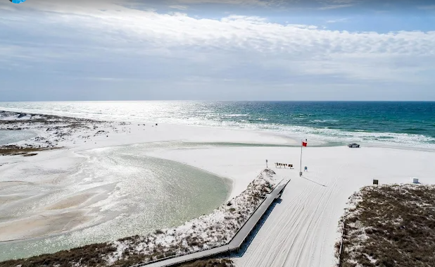 Beach with sand dunes, a small ramp, and a red and white flag, with the ocean in the background under a cloudy sky.