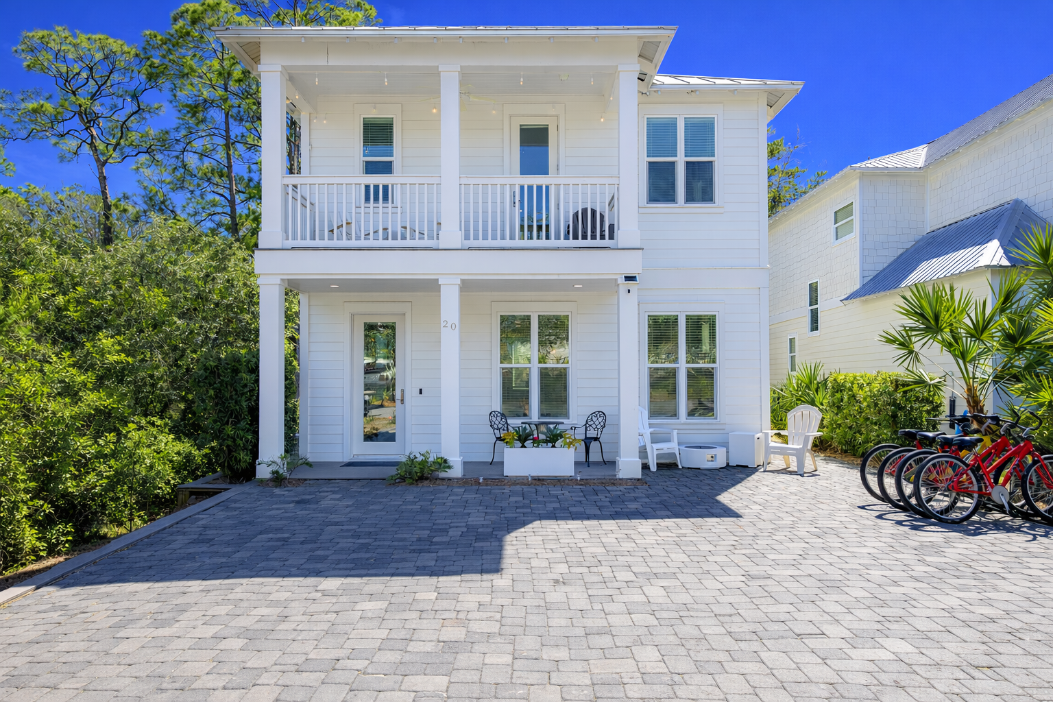 Second Set exterior with a bright white coastal facade, covered front porch, upper balcony, lush landscaping, and beach cruiser bikes.