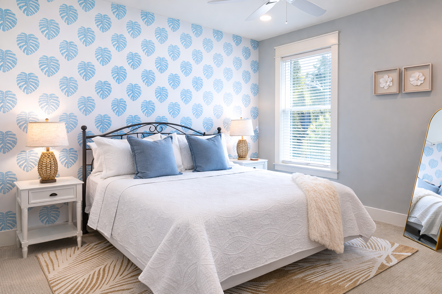 Bedroom with white bedspread, blue pillows, and tropical leaf wallpaper; white nightstands with lamps, window with blinds, and a large mirror leaning against the wall.