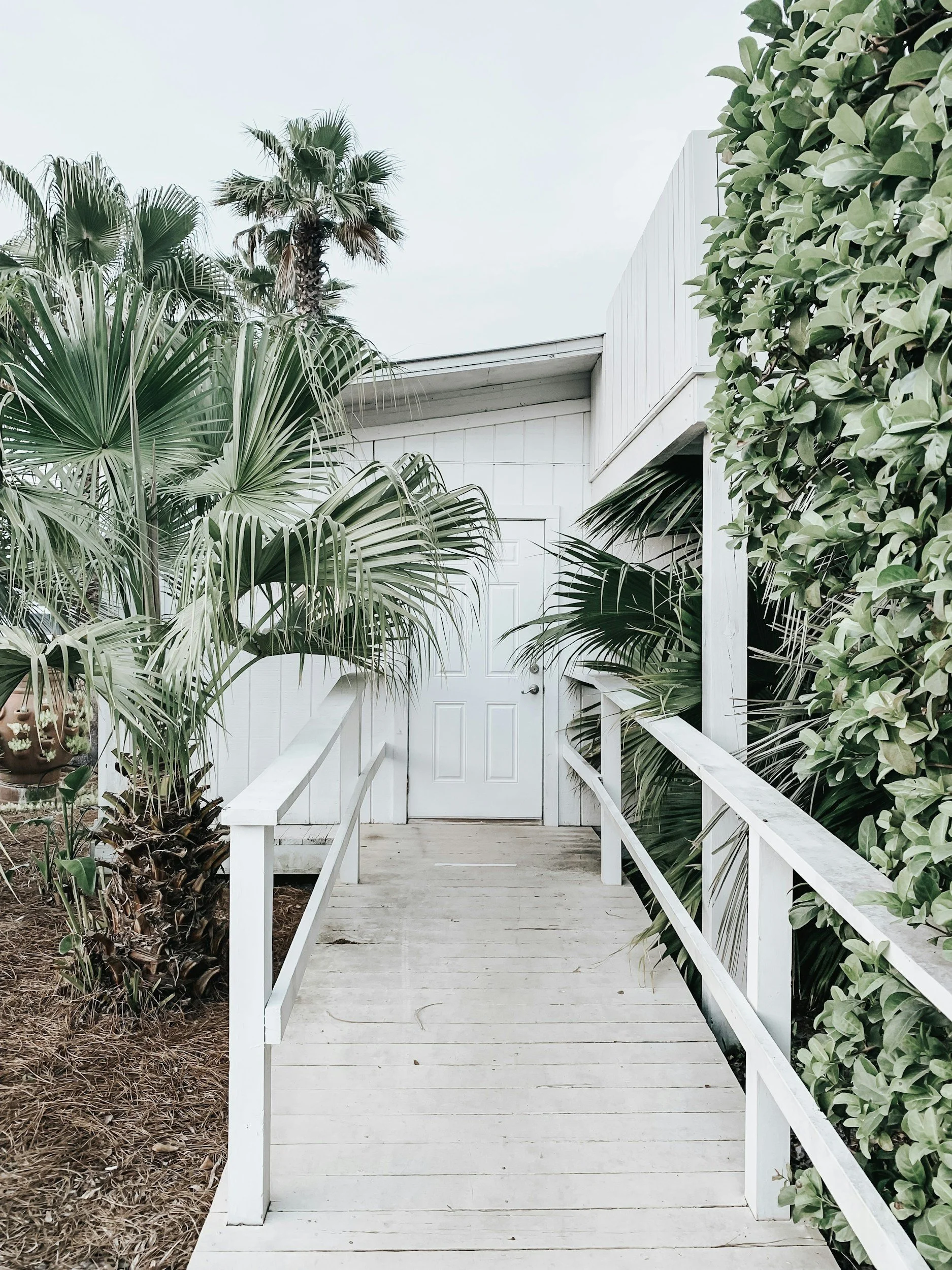 White wooden door at the end of a small wooden bridge with white railings, surrounded by green tropical plants.