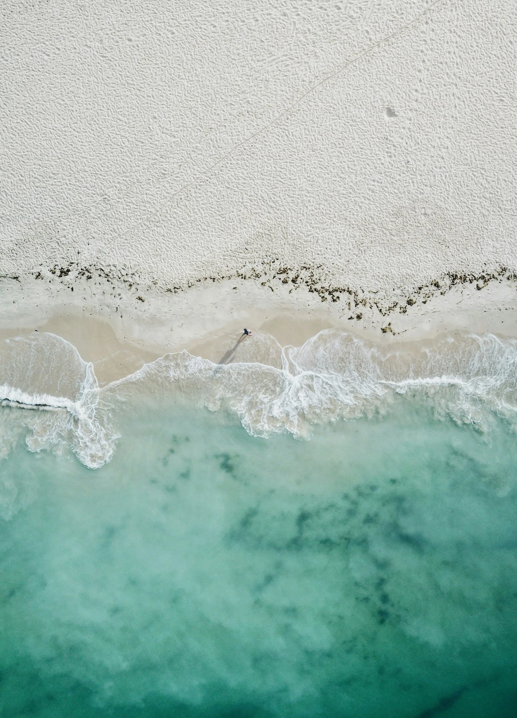Aerial view of a beach with white sand, gentle waves, and a person walking along the shoreline.