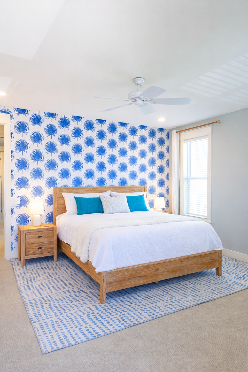 A bedroom with a wooden bed frame, white bedding, blue and white pillows, a blue patterned accent wall, two bedside tables with lamps, a patterned rug, a window with white curtains, white ceiling fan, and recessed lighting.