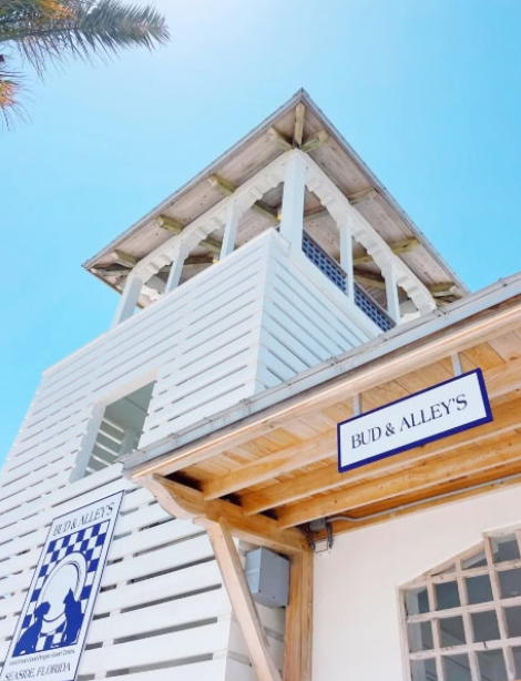 A white wooden building with a tower on top, featuring a sign that reads 'BUD & ALLEY'S' and another sign with a blue and white boat logo, set against a clear blue sky.