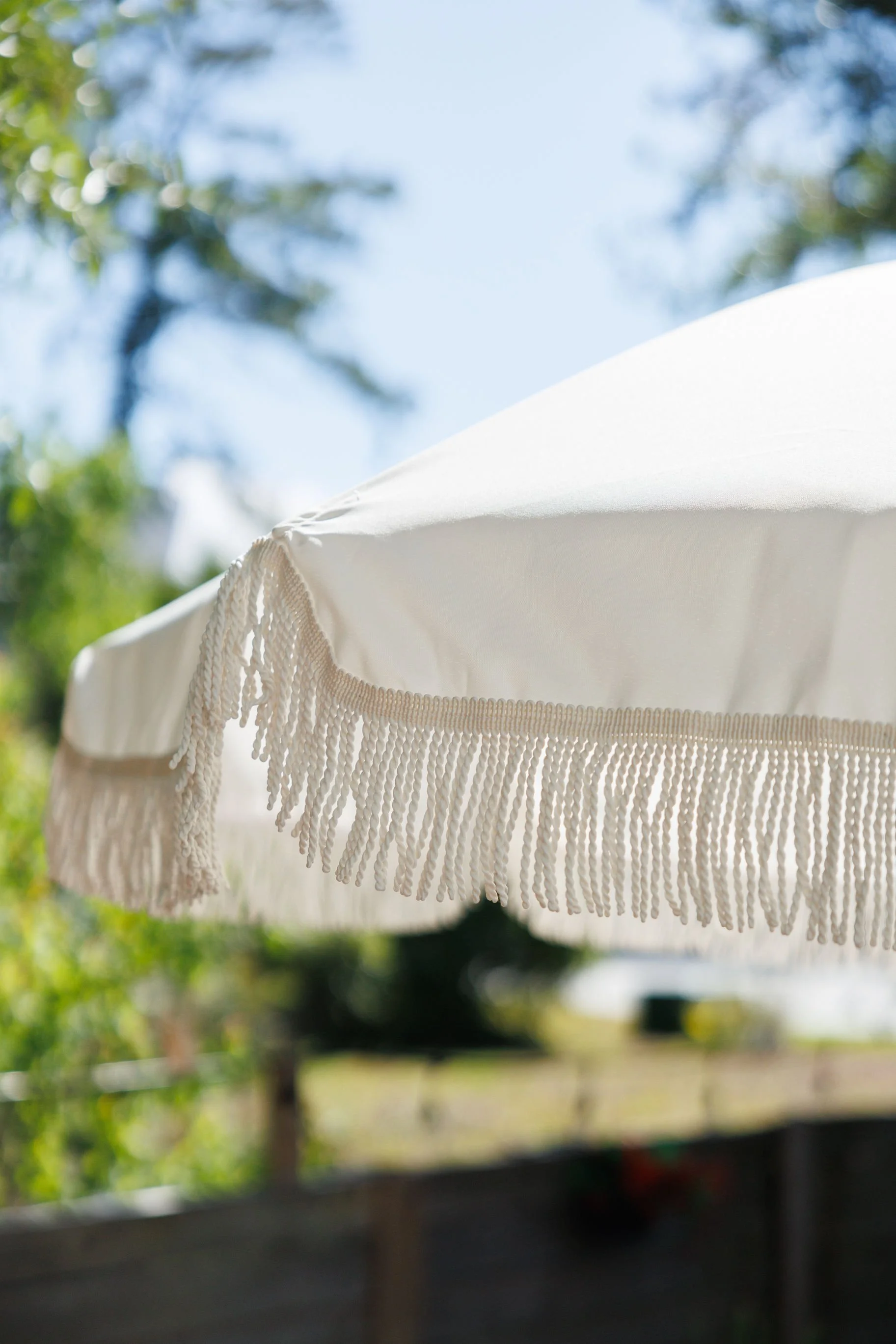 Close-up of a white outdoor patio umbrella with decorative fringes, against a bright blue sky and blurred greenery in the background.