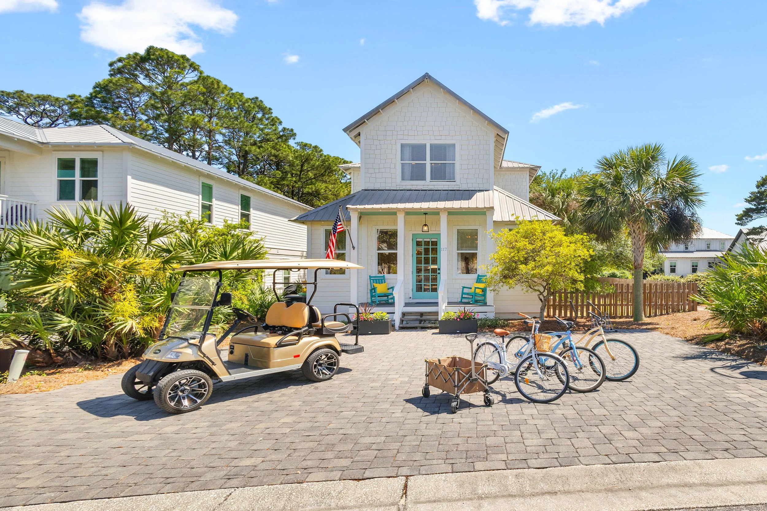 A white residential house with a small front porch, blue chairs, palm trees, and multiple bicycles and a golf cart parked on a brick driveway under a clear blue sky.