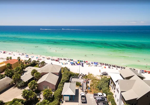 View of a beach with turquoise water, white sand, umbrellas, and people. Residential buildings with garages and greenery in the foreground. Boats on the water in the distance.