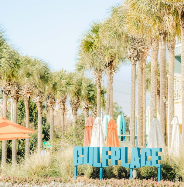Beachside scene with tall palm trees, colorful umbrellas, and a blue sign reading 'GULF PLACE' in front of the sand dunes.