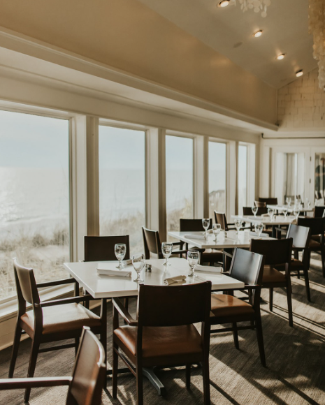 Dining area with tables set for a meal, featuring wine glasses and silverware, near large windows with a view of outdoors.