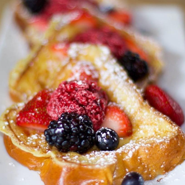 Close-up of a French toast topped with mixed berries, sliced strawberries, powdered sugar, and syrup.