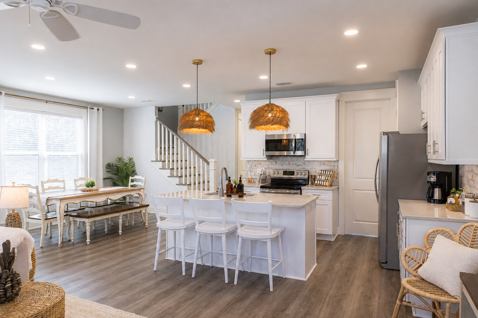 Bright, open-concept kitchen and dining area with white cabinetry, stainless steel appliances, a kitchen island with barstools, and a large window with white curtains. Decor includes woven pendant lights, a potted plant, and wicker chairs.