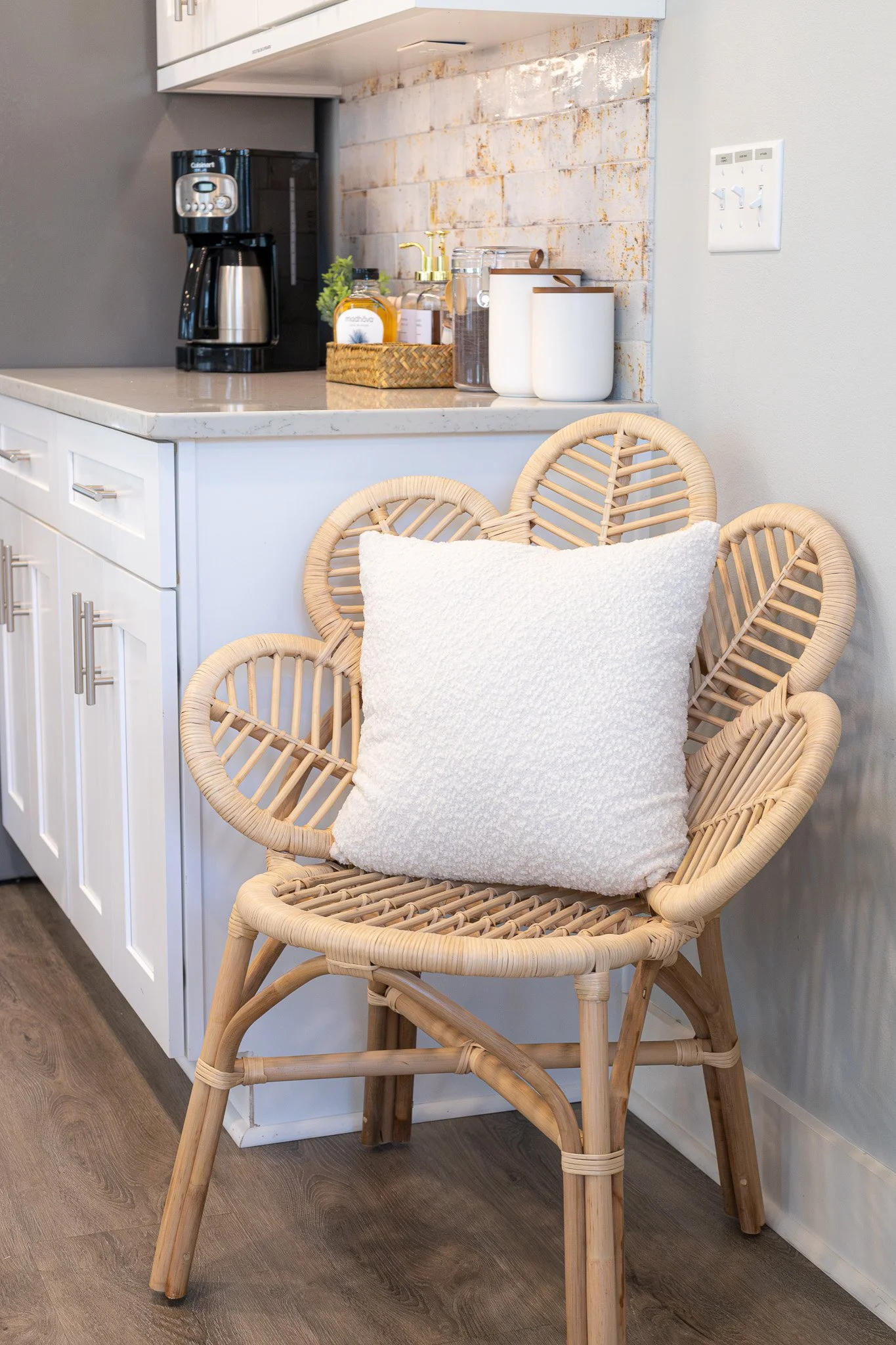A wicker chair with a white fluffy pillow resting on it, positioned in a kitchen corner next to a white cabinet with a marble countertop. The kitchen includes a coffee maker, soap dispenser, and storage jars on the counter, with a brick accent wall behind.