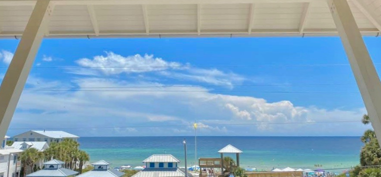 View from a balcony overlooking a beach with blue sky, ocean, and clouds, with beach houses and umbrellas in the foreground.