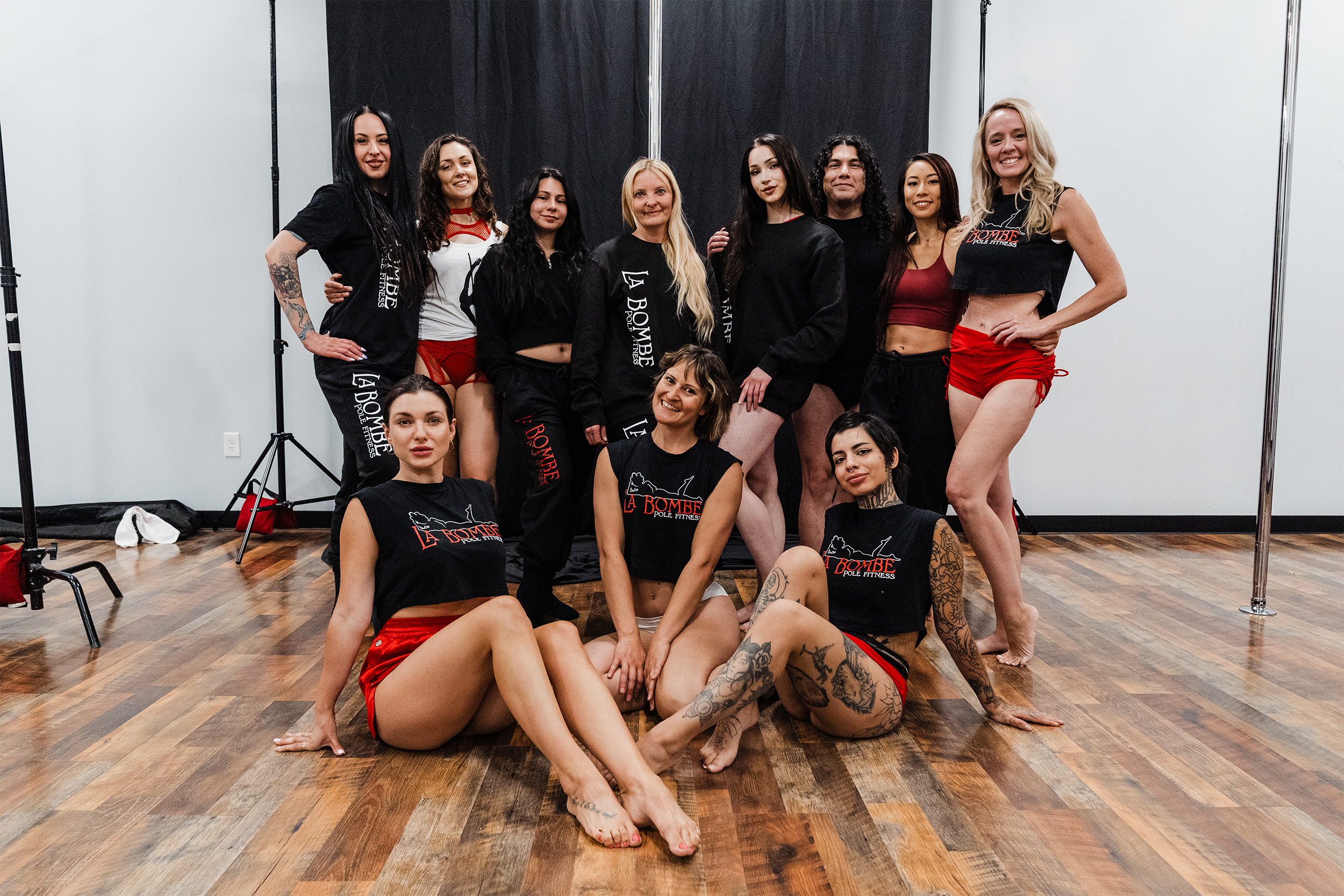 Group of women in athletic wear posing in a fitness studio with wooden floors, black curtains, and studio equipment.