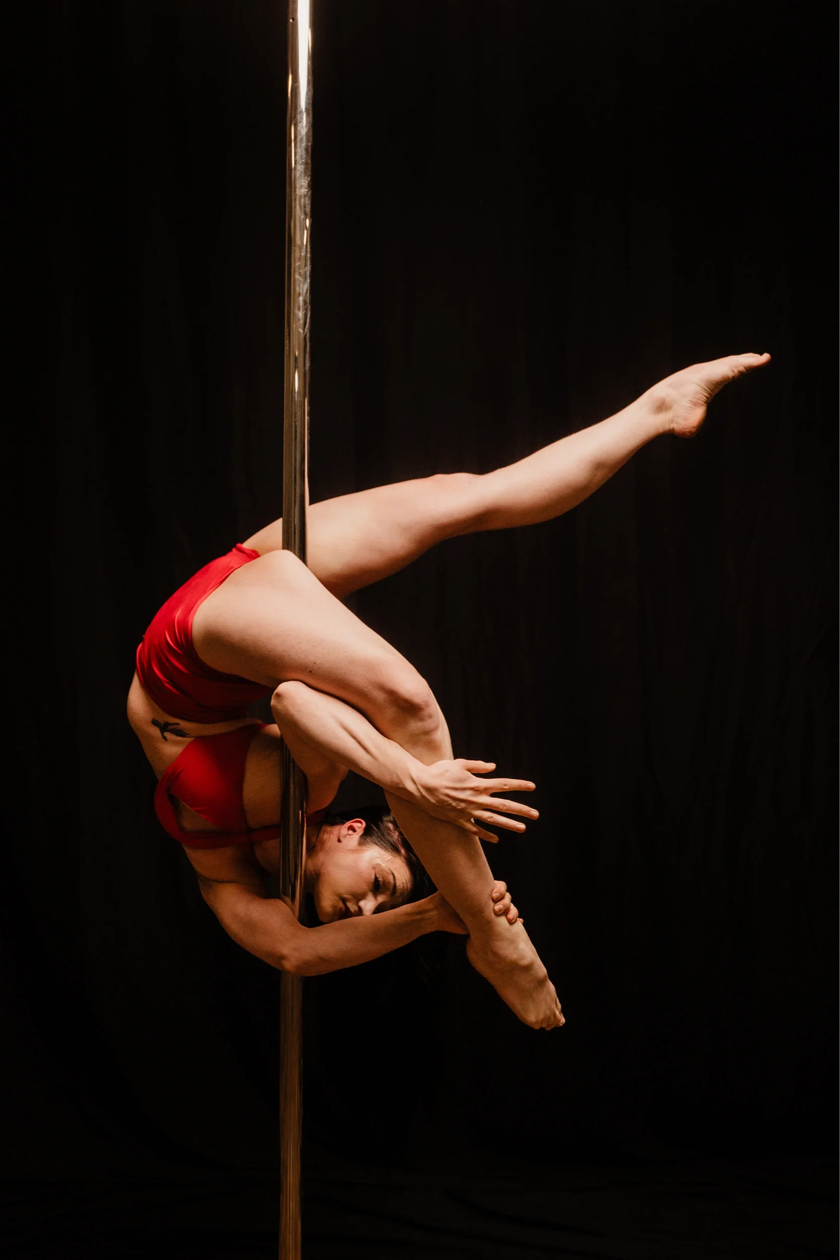 A woman performing a pole dance upside down against a black background, dressed in red athletic wear, gripping the pole with her legs and one arm while her other arm is extended.