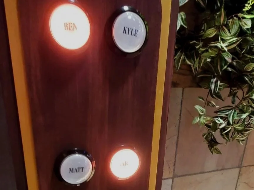 Wooden wall with four circular lights labeled with names: Ben, Kyle, Matt, and Air. Two lights are illuminated, two are off. To the right, a potted plant with green and cream-colored leaves.