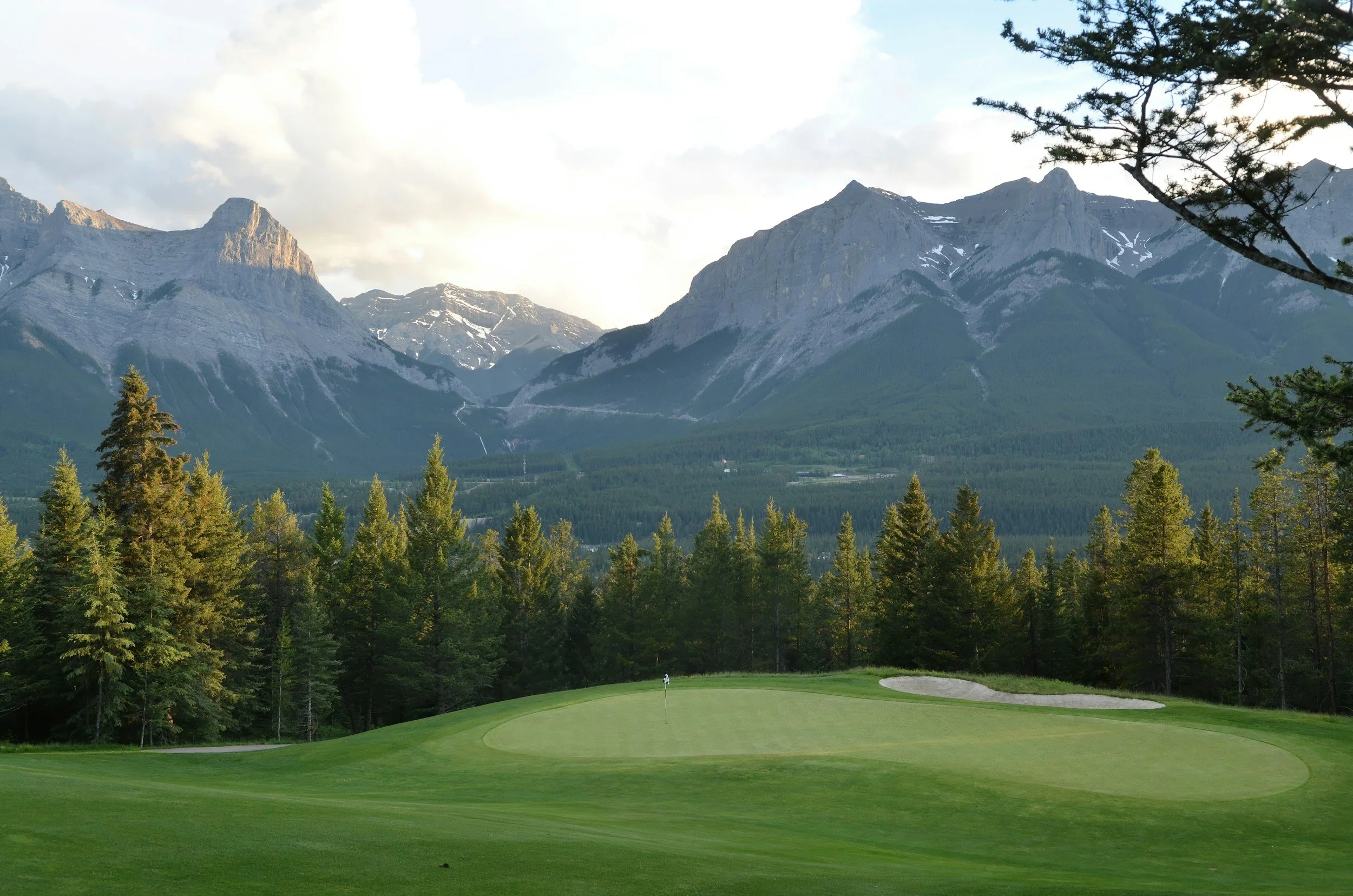 A golf green with a flag and sand trap, surrounded by a forest of tall pine trees, with mountains in the background and a partly cloudy sky.