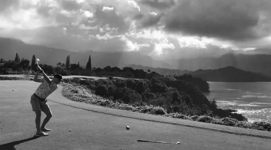 Person playing golf on a golf course near the coast, with water and mountains in the background, under cloudy skies.