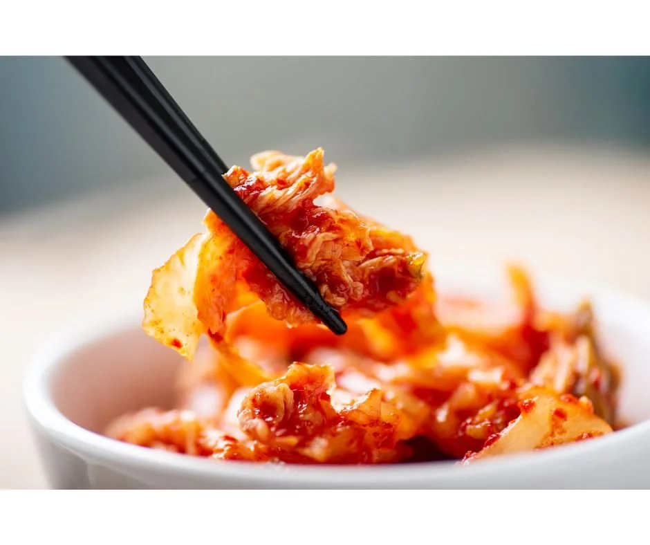 Close-up of chopsticks holding a piece of kimchi over a white bowl filled with kimchi.