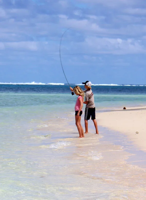 Two children, a boy and a girl, stand at the shoreline of the beach, with one child fishing into the ocean.