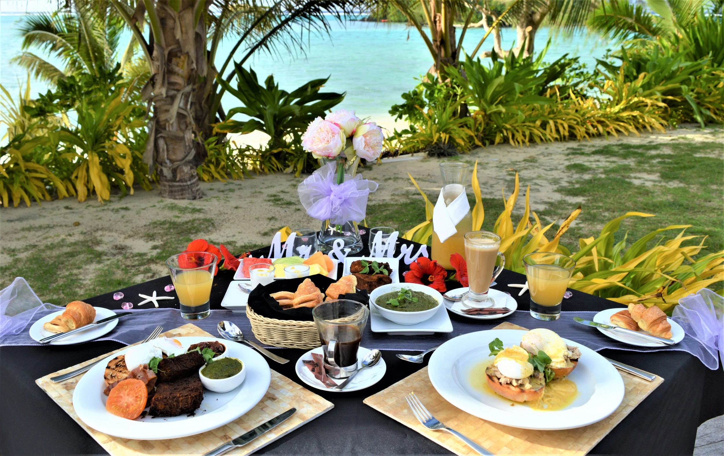 A table set for a meal outdoors on a tropical beach, with a variety of dishes including salads, pastries, and drinks, decorated with flowers and 'Mr. & Mrs.' signs.