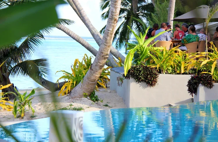 A group of people dining at an outdoor restaurant by the beach, with palm trees and tropical plants nearby.